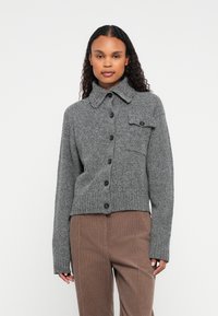 Kobieta w szarej dzianinowej kardiganie z guzikami i kieszonką na piersi, stojąca na tle białej ściany. Ma na sobie brązowe spodnie.