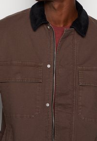 Man wearing brown jacket with black corduroy collar and zipper over a red crew neck shirt, cropped at the neck and waist.