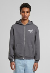 Grå zip-up hettegenser laget av en myk bomullsblanding, med en sommerfuglgrafikk på venstre bryst og frontlommer.