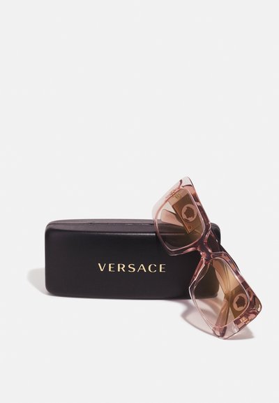Óculos de sol Versace com armação transparente rosa e lentes castanhas degradê, apoiados numa caixa preta com logótipo dourado.
