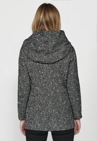 Čierno-biely kabát s flekovaným vzorom a štruktúrovaným povrchom, s kapucňou a dlhými rukávmi, so štíhlym strihom a strednou dĺžkou.