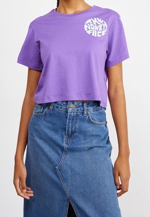 Kobieta w fioletowym, krótkim t-shircie z logo The North Face oraz niebieskiej dżinsowej spódnicy z wysokim stanem i przednim rozcięciem, z rękami swobodnie opuszczonymi wzdłuż ciała.