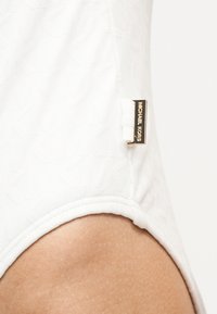 Κοντινή λήψη λευκού υφασμάτινου υφάσματος με χρυσή ετικέτα λογότυπου Michael Kors στη ραφή στο πλευρό κοντά στον γοφό ενός ατόμου.