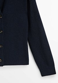 Manche de cardigan en maille côtelée bleu marine et devant boutonné sur fond blanc, montrant la texture et les détails des boutons.