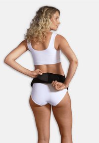 Musta vyötärön tuki, joka on puettu valkoisen paidan ja alushousujen päälle, esittelee sileää, elastista materiaalia ja ergonomista muotoilua selkätuen tarjoamiseksi.