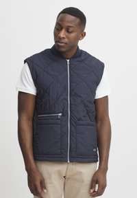 Marinblå quiltad väst med dragkedja framtill, hög krage och två framfickor, varav en med dragkedja. Slät textur med en lätt design.