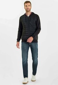 Felpa nera con pannelli anteriori trapuntati e colletto a contrasto bianco e rosso. Abbinata a jeans blu e sneaker bianche su uno sfondo neutro.