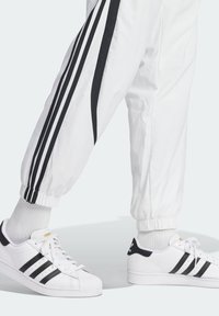 Vita idrottsbyxor med svarta sidostråkar och elastiska mudd, kombinerade med vita sneakers med tre svarta ränder och en strukturerad tåhuvud.