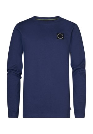 Person iført en blå langærmet skjorte med en lille sort logopatch, lyseblå opsmøgede jeans og hvide sneakers, der står mod en hvid baggrund.