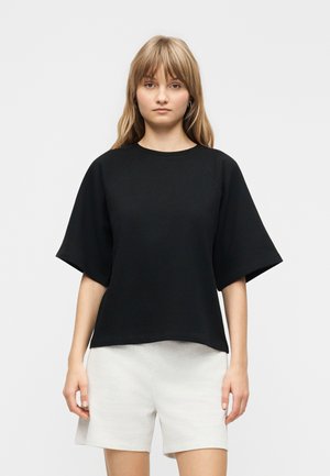 Γυναίκα με φαρδιά μαύρη κοντομάνικη μπλούζα και άσπρο σορτς στέκεται μπροστά σε ένα απλό ανοιχτό γκρι φόντο.