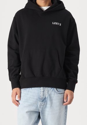 Mann trägt schwarzen Levi’s Hoodie mit Fronttasche und hellblauen Jeans vor einfarbigem Hintergrund.