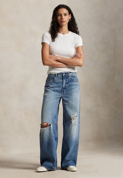 Vide bukser i denim med slitte detaljer i lys blå, med frynsete revner. Kombinert med en tettsittende hvit bomulls-T-skjorte og hvite joggesko.