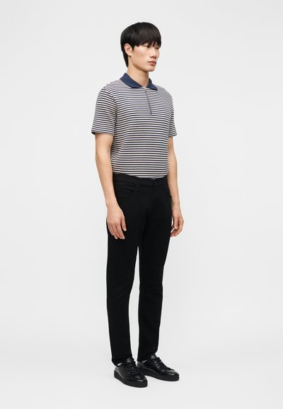 Noor mees seisab, kandes triibulist lühikese varrukaga polo särki, musti pükse ja musti tossusid, taustal on tavaline valge taust.