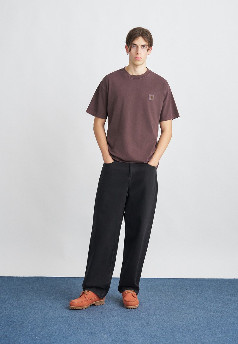 Braunes Baumwoll-T-Shirt mit kleinem Logo, lockere schwarze Jeanshosen und orangefarbene Ledersandalen mit strukturierten Sohlen. Einfaches, lässiges Outfit.
