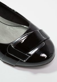 Chaussure plate en cuir verni noir avec un détail de boucle rectangulaire argentée brillante sur l’orteil arrondi.