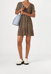 Schwarz mit Blumenmuster, Kleid mit kurzen Ärmeln, V-Ausschnitt und geraffter Taille. Kombiniert mit einer hellblauen Handtasche und klobigen weißen Schuhen.