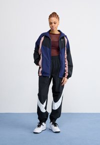 Giacca a vento Nike in blu navy, rosa e nero con pannelli a contrasto; abbinata a pantaloni neri con grandi loghi bianchi; sneaker bianche.