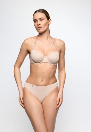 Donna che indossa un reggiseno beige con spalline decorative ad anello e biancheria intima abbinata, in piedi davanti a uno sfondo chiaro e uniforme.