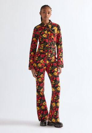 Nő, aki egy testhezálló, virágos mintájú kezeslábasban áll, amelyen piros, sárga és zöld virágok vannak, egy egyszerű fehér háttér előtt.