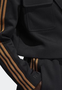 Černá sportovní bunda se zlatými pruhy. Má strukturovanou tkaninu, přední kapsu a vyšívaný detail na manžetě.
