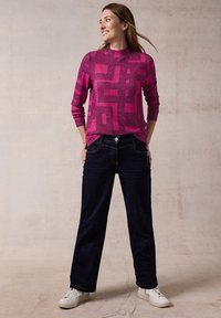 Fuchsia trui met lange mouwen en geometrisch patroon, gecombineerd met donkerblauwe brede jeans en witte sneakers. Zacht, gestructureerd materiaal.