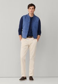 Man staat met handen in de zakken, draagt een blauw gewatteerd vest, een marineblauw shirt met lange mouwen, beige broek, bruine riem en bruine loafers tegen een grijze achtergrond.