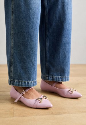 Person iført blå jeans og lyserøde lakflade sko med guldbeklædt spænde, stående på trægulv.