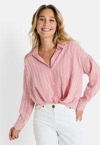Camisa de botones a rayas rojas y blancas con mangas largas, corte relajado y detalle de nudo en la parte delantera, combinada con pantalones blancos de cintura alta.