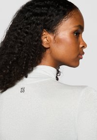 Weißes Langarmoberteil mit hohem Kragen und strukturiertem, schimmerndem Stoff. Verfügt über ein schwarzes "HUGO"-Logo hinten in der Nähe des Kragens.