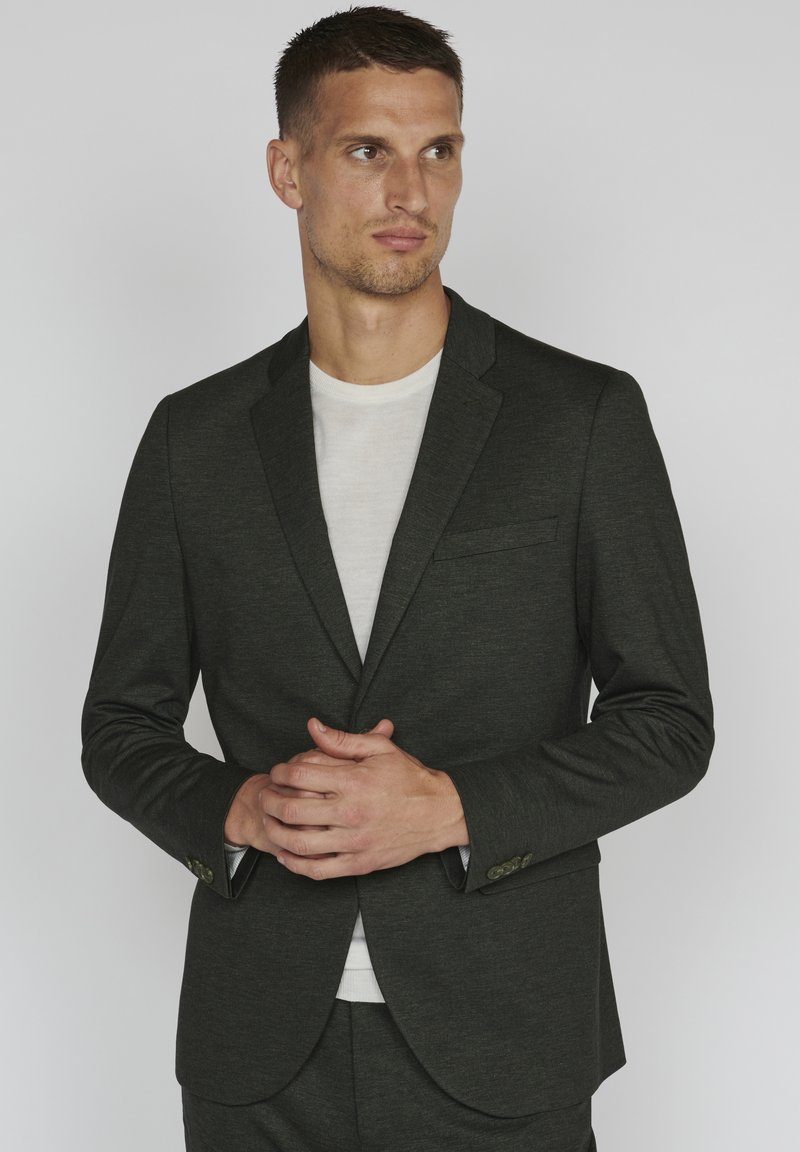 Dark green textured blazer with a single chest pocket and buttoned cuffs, paired with a fitted white shirt, displayed against a light background.
