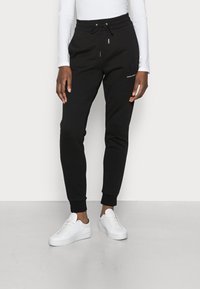 Svarta träningsoverallsbyxor i mjukt tyg, med dragsko i midjan, avsmalnande ben, ribbade mudd och sidofickor. Tillsammans med vita sneakers.