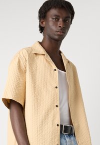 Short-sleeve, textured shirt in light yellow with a turn-down collar, featuring black buttons and worn over a white tank top and denim.