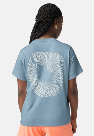 Persona con el cabello trenzado que lleva una camiseta azul claro con texto blanco en espiral en la espalda y pantalones cortos color melocotón, de pie frente a un fondo liso.