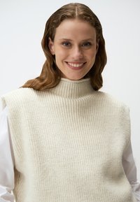 Gilet en tricot de couleur crème avec un col haut, texture côtelée et sans manches, porté par-dessus une chemise blanche à manches longues.