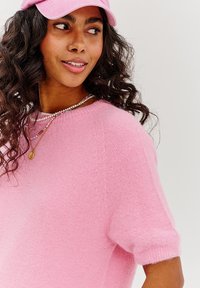 Ružový oversized pletený sveter s krátkymi rukávmi, ktorý má jemnú textúru a zaoblený výstrih, doplnený o delikátne náhrdelníky.