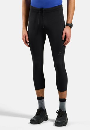 Mann trägt schwarze dreiviertellange Sportleggings, graue Sportsocken, schwarze Laufschuhe und ein blaues Shirt und steht vor einem weißen Hintergrund.