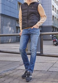 Czarna kamizelka puchowa na beżowym hoodie, niebieskie dżinsy i czarne sneakersy. Kamizelka ma zamek błyskawiczny z przodu i detal z logo. Miejski krajobraz.