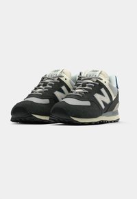 A New Balance 574 sneaker fekete és szürke velúr felső résszel, krémszínű kiegészítőkkel és textúrázott gumitalppal rendelkezik, kék sarokrészlettel.