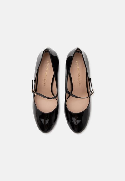 Paire de chaussures Mary Jane en cuir verni noir avec fines brides et boucles dorées sur fond blanc.