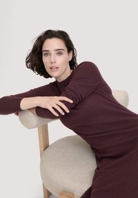 Burgunderfarbendes Strickkleid mit hohem Kragen und langen Ärmeln, kombiniert mit einem Stuhl aus leichtem Material mit Holzrahmen und runder Sitzfläche.