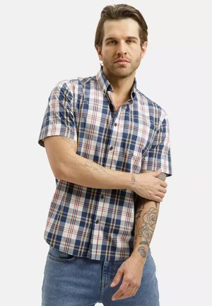 Mężczyzna w niebiesko-biało-czerwonej koszuli w kratę z krótkim rękawem, pokazujący wytatuowane ramiona, poprawia rękaw, stoi na prostym tle.