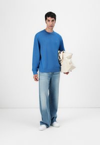 Maglione blu con maniche lunghe, collo rotondo, abbinato a jeans a gamba larga blu chiaro. Porta una borsa a tracolla beige. Tessuto morbido, design casual.