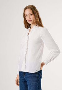 Giovane donna con capelli mossi, che indossa una blusa bianca con volant e jeans blu, in piedi su uno sfondo chiaro e semplice.