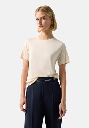 Blonde vrouw die een roomkleurige top met korte mouwen draagt, ingestopt in een hooggetailleerde donkerblauwe broek met een zichtbaar logobandje.