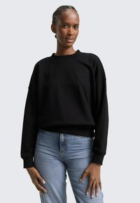 Donna che indossa una felpa nera a maniche lunghe con testo sottile e jeans azzurri, in piedi con le mani rilassate lungo i fianchi.