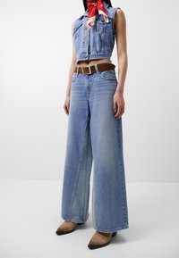 Svetle modr�ý džínový cropped vest se zapínáním na knoflíky, spárovaný s širokými světle modrými džíny. Obsahuje hnědý opasek a béžové lodičky na podpatku.