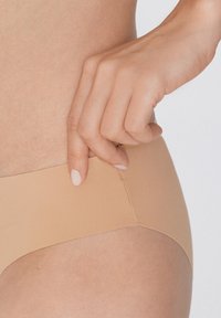 Nude bezešvé spodní prádlo s hladkou texturou, nízkým pasem a jemnými švy, drženo rukou na neutrálním pozadí.