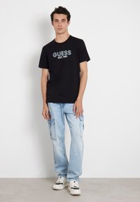 Camiseta negra de algodón con el gráfico "GUESS EST. 1981", combinada con pantalones cargo de color azul claro y zapatillas negras y blancas. De pie contra un fondo neutral.