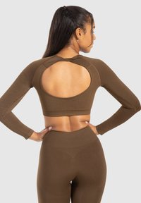 Donna che indossa un crop top a maniche lunghe marrone aderente con un grande taglio ovale sulla schiena e leggings a vita alta coordinati, con le mani sui fianchi.