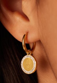 Gouden ringoorbel met een ronde hanger met een handontwerp, omringd door heldere kristallen. Geleefde oppervlakte en glanzende afwerking.